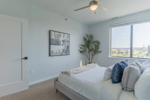 A bedroom in a River Rock residence with a platform bed, an olive tree, and a window over the rooftops of Mishawaka.